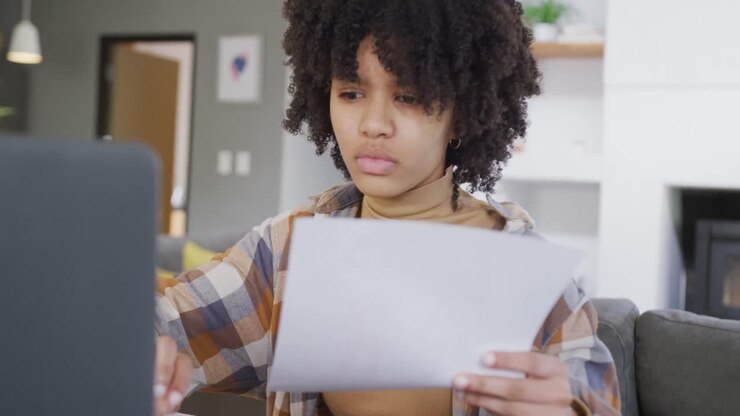 African american teenage girl reading paperwork and using laptop at home, slow motion