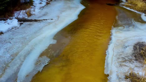 mineral river thaw frosty iced shore reveal caves under stone bridges yellow tall grass forest snow banks dolly roll aerial drone overlooking dense bushes trees ice tips of streaming waters