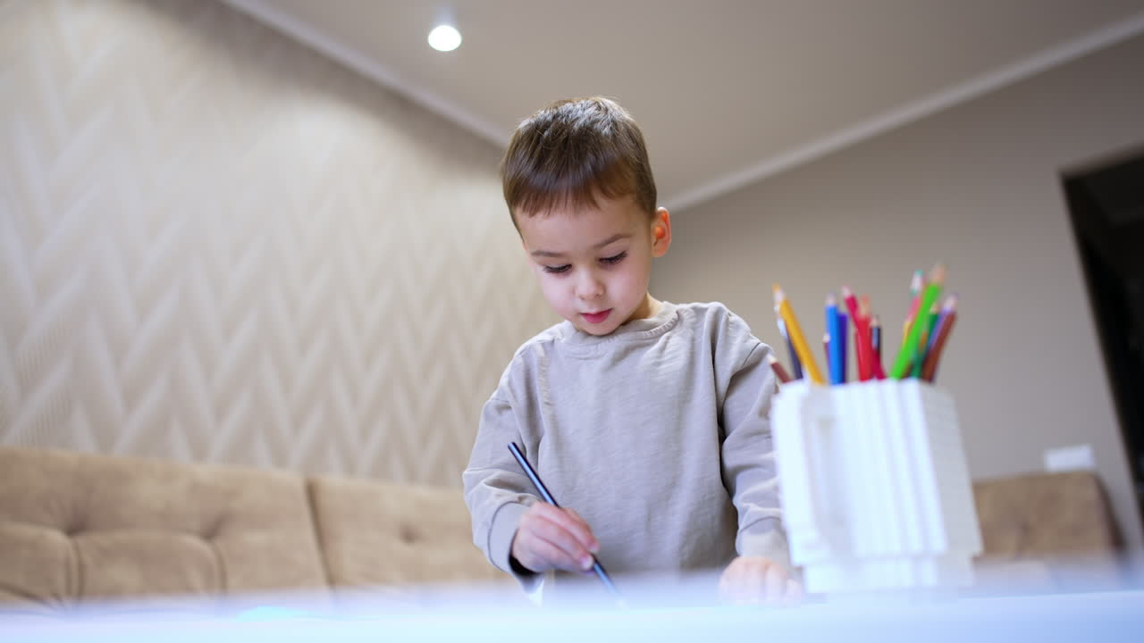 Adorable Caucasian toddler draws sitting at desk. Low angle view at the baby boy doing art.