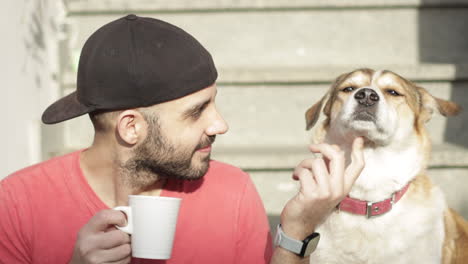 caucasian man drinks a cup of tea for break or breakfast next to his brown and white or red dog