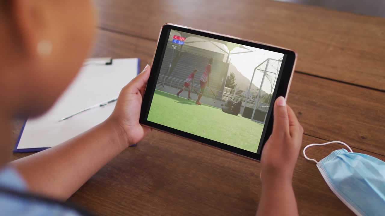 태블릿에서 하키 경기를 시청하는 얼굴 마스크와 함께 테이블에 앉아있는 여성의 복합