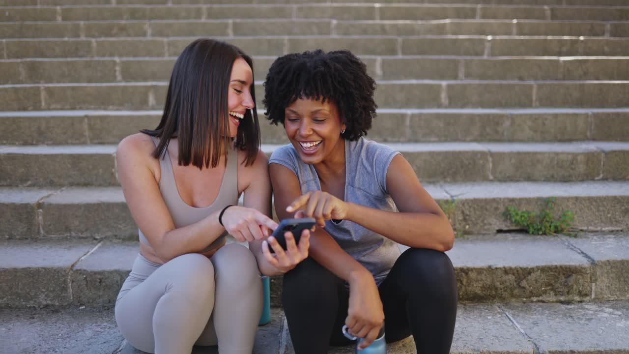 Two Happy Young Women Laughing and Sharing a Smartphone on Stone Steps