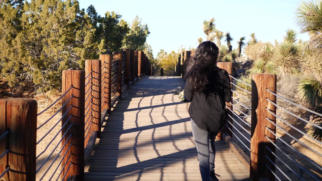 una chica con el pelo largo y negro cruza un puente de madera en el desierto a cámara lenta
