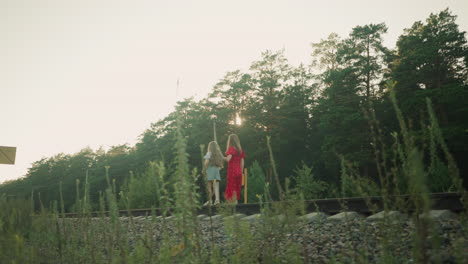 back view of little girl walking carefully on rail beam while older companion in red dress gently holds her hand as they interact along forest railway path under soft evening