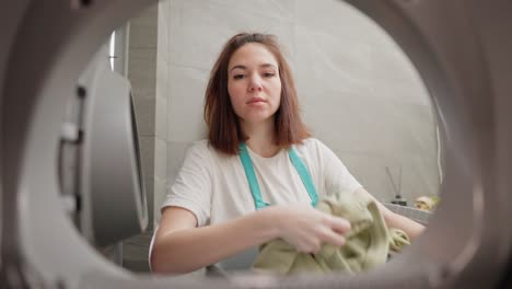 vista desde el interior de la lavadora una confiada chica morena con una camiseta blanca y un delantal azul carga cosas sucias en la lavador de un lavabo de plástico gris y luego lo enciende