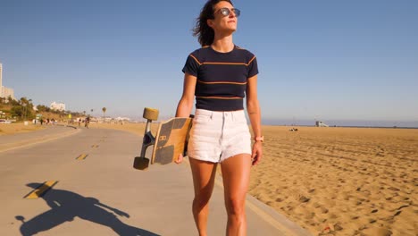 girl in sunglasses carrying longboard while walking at the skatepark in summer - slow motion