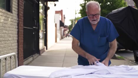 an elderly man in scrubs skillfully folding white sheets on a table outside, demonstrating care and attention to detail in his task