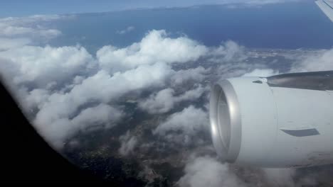 a first-person point-of-view shot from inside the cabin of a flying airplane, gazing out the window at one of the wing engines as it traverses through sparse clouds