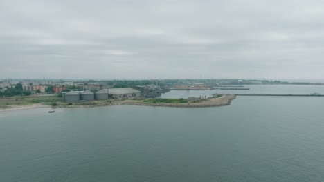 Vista-Aérea-De-Grúas-Portuarias-Y-Muelles-De-Carga-Vacíos-En-El-Puerto-De-Liepaja,-Ciudad-De-Liepaja-En-El-Fondo,-Día-De-Verano-Nublado,-Amplia-Toma-De-Drones-Avanzando