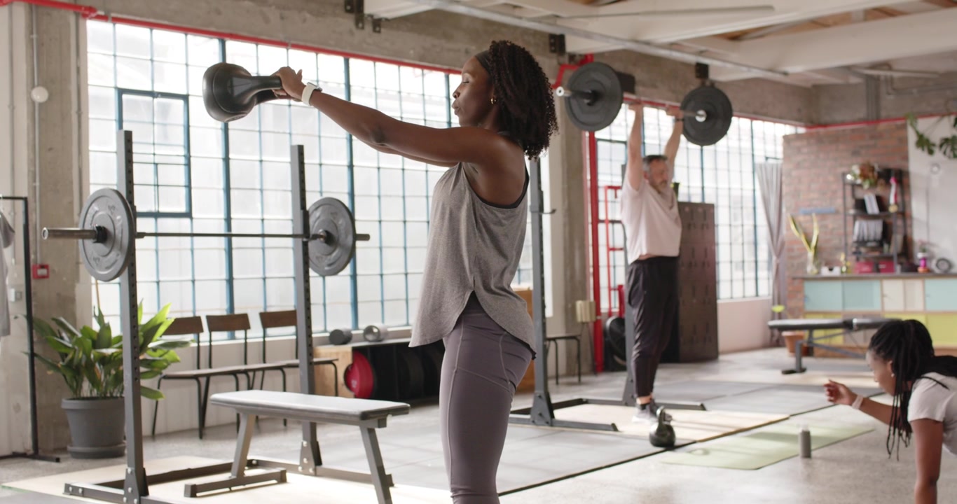 Diverse trio hinging hip swinging kettlebell pressing barbell reaching plank, training in gym gear