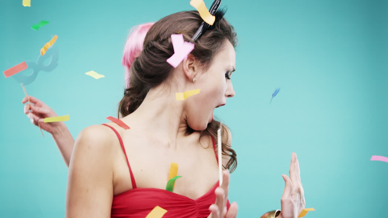 빨간 드레스와 분홍색 머리카락을 입은 여자 친구들이 느린 움직임의 파티 사진 부스에서 춤추고 있습니다.