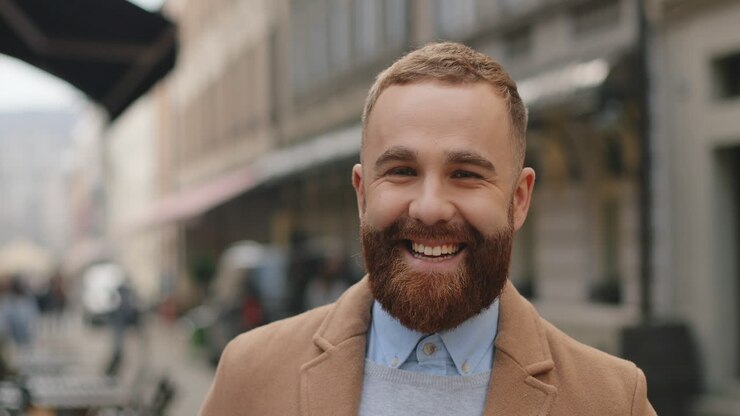 Close-up view of caucasian cheerful businessman with beard in elegant clothes looking at the camera and smiling on the street