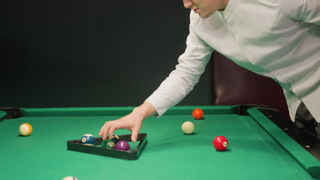 billiard player spot picking billiard balls on green pool table, placing them in triangle rack. focused on precise arrangement for upcoming game in dimly lit indoor setting, preparing for break shot