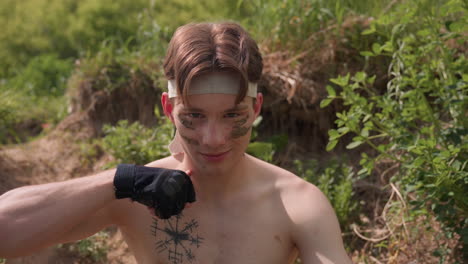 shirtless soldier warrior in bandage headband with muddy camouflage stripes, sitting amidst green brown foliage on eroded hillside, eyes fixed on camera, subtle smile and calm gaze