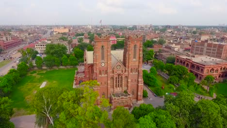 Luftaufnahme-Einer-Schönen-Alten-Kirche-Mit-Stadt-Im-Hintergrund,-Schöne-Bäume-Und-Gras-Rund-Um-Die-Kirche,-Tauben,-Die-über-Die-Kirche-Fliegen,-Menschen,-Die-Sich-Außerhalb-Der-Kirche-Bewegen