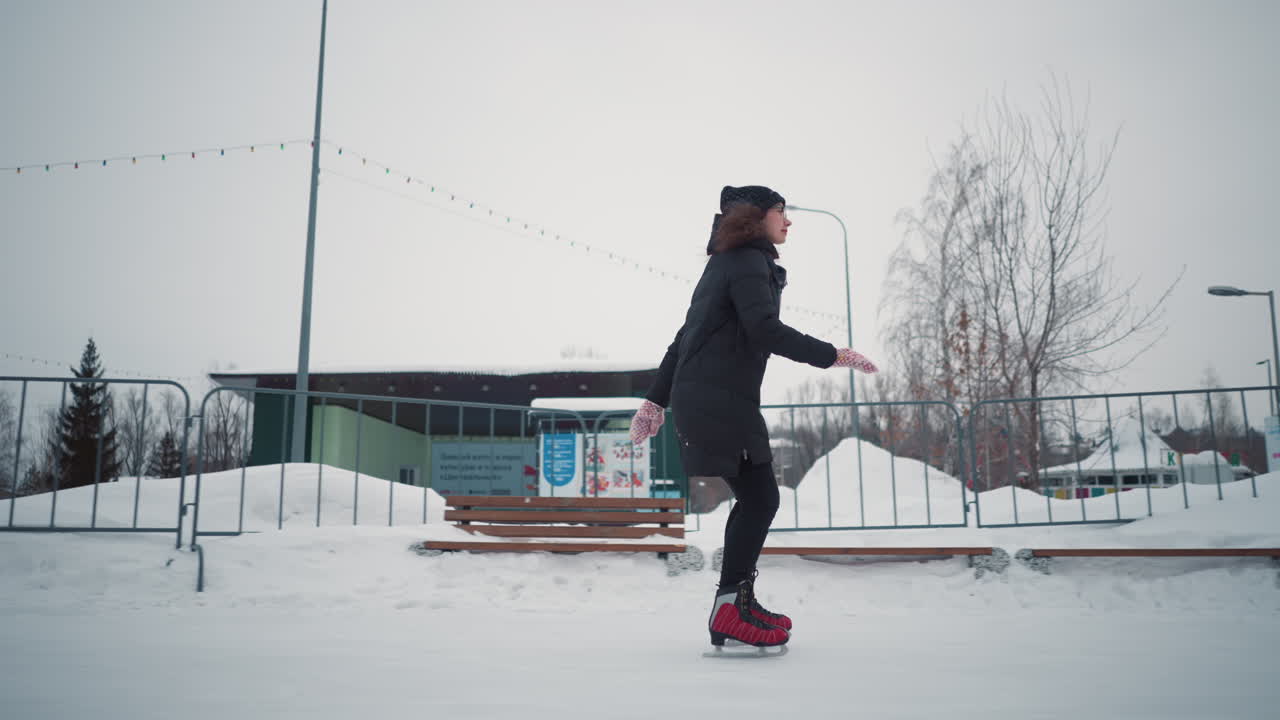 Lady skating on outdoor ice rink in winter wearing black coat, red skates, scarf, gloves, and hat, maintaining balance gracefully while snow, benches, metal railings, and trees create seasonal background