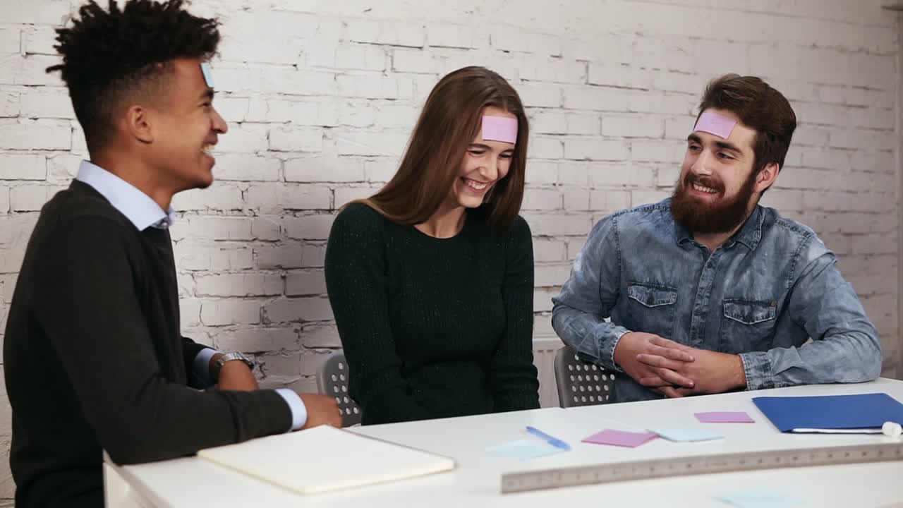 jóvenes hermosos en la oficina sentados en la mesa jugando al juego de nombres con notas adhesivas pegadas a su frente