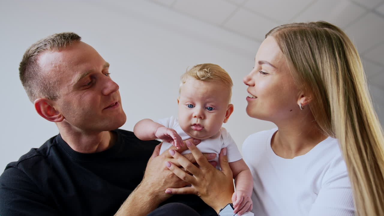 Happy smiling Caucasian people holding the cute baby son. Loving parents kiss their blue-eyed infant on the cheek.