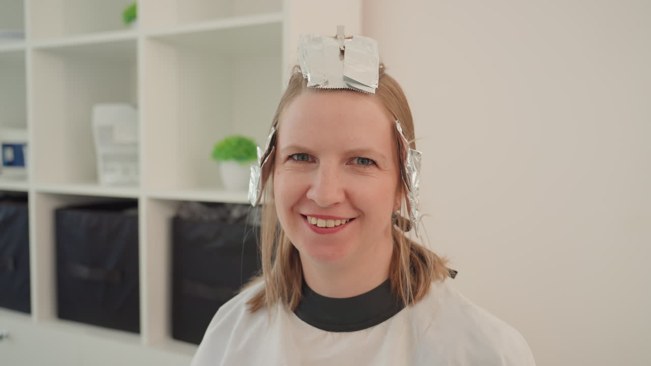 smiling woman in salon with foil, beaming as stylist checks color and adjusts foils, protective cape worn, modern shelving and potted plant in background, bright warm lighting, candid joyful makeover