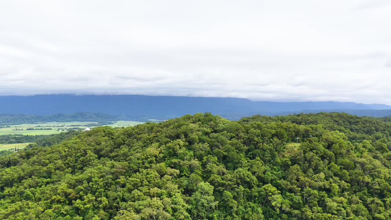Drone glides over dense green rainforest hill, revealing distant mountains under cloudy daylight