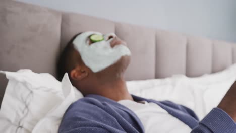 Video-of-african-american-man-lying-on-bed-with-cleansing-face-mask-and-cucumber-on-eyes-in-bedroom