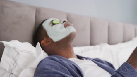 video of african american man lying on bed with cleansing face mask and cucumber on eyes in bedroom