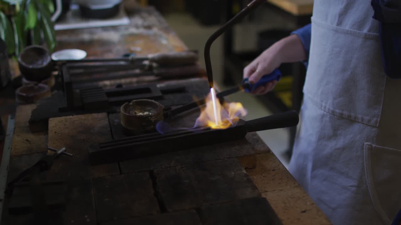 Midsection of caucasian female jeweller in workshop wearing apron, using gas burner