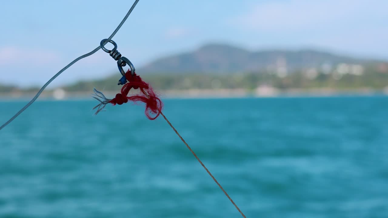 A fishing line with a red knot sways gently over the ocean, set against a backdrop of distant hills in Phuket