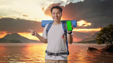 excursionista masculino asiático con mochila de montañismo que muestra una maqueta de pantalla verde de un teléfono inteligente y apunta hacia un lado en un lago