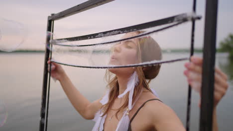 una joven artista sopla muchas burbujas de jabón muestra un espectáculo teatral usando un marco en cámara lenta al atardecer en un lago