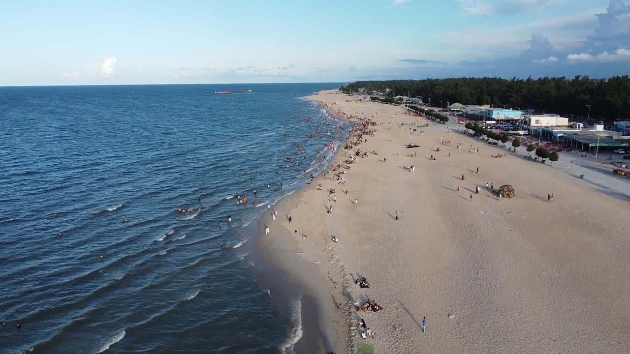 Drone footage of Nha Trang's bustling beach captures people enjoying vibrant coastal leisure under bright skies. The sandy shoreline meets azure waters in a dynamic aerial view