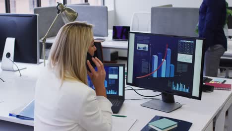 Caucasian-businesswoman-sitting-at-desk-using-computer-talking-on-smartphone-in-office