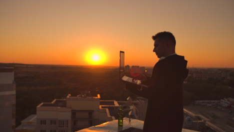 Hacker-using-laptop-on-rooftop-with-city-view-and-forex-chart.-Hacking-and-stats-concept.-A-man-at-sunset-in-slow-motion-writing-software-code-on-a-laptop