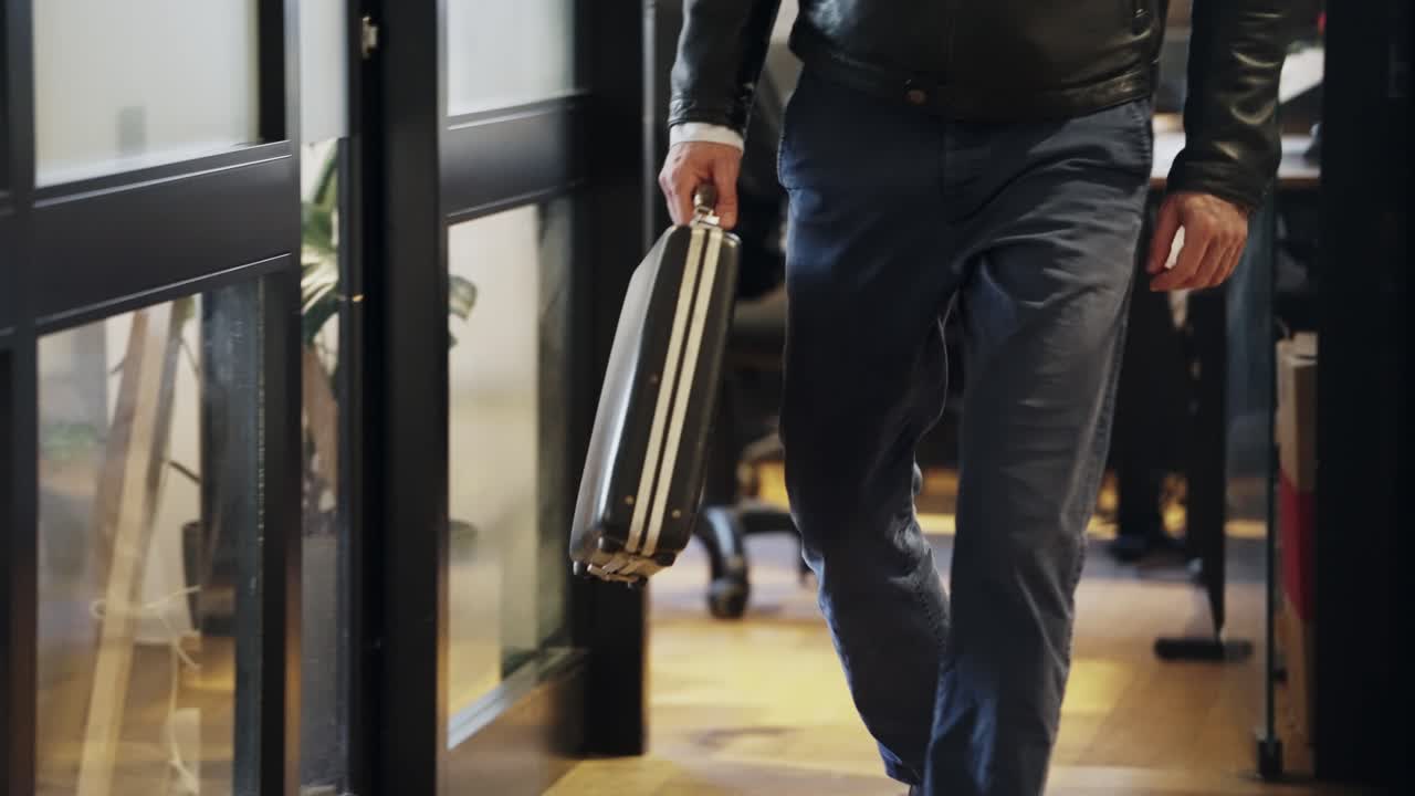 Man walking with a black briefcase in hand, wearing a leather jacket and jeans. Business, travel, secret deal, corporate, negotiation, undercover operation, crime, finance, departure, or investigation