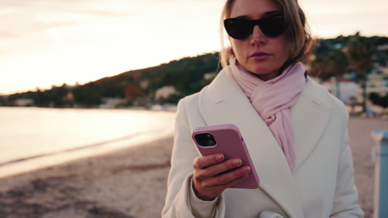 Donna elegante con cappotto bianco, sciarpa e occhiali da sole che controlla il telefono mentre è in piedi su una spiaggia al tramonto