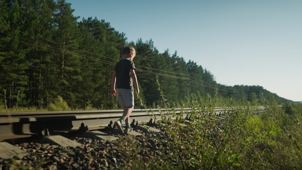 Little kid wearing dark shirt and gray shorts walks carefully along rail track in forest as sunlight creates soft glow in background, with power lines and trees framing quiet, peaceful countryside