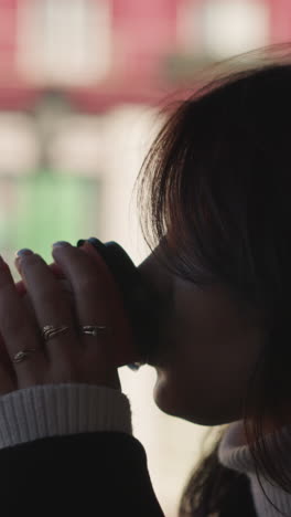 primer plano de una mujer bebiendo café de una taza al aire libre, dedos adornados con anillos, edificio rojo borroso y luz natural suave en el fondo creando