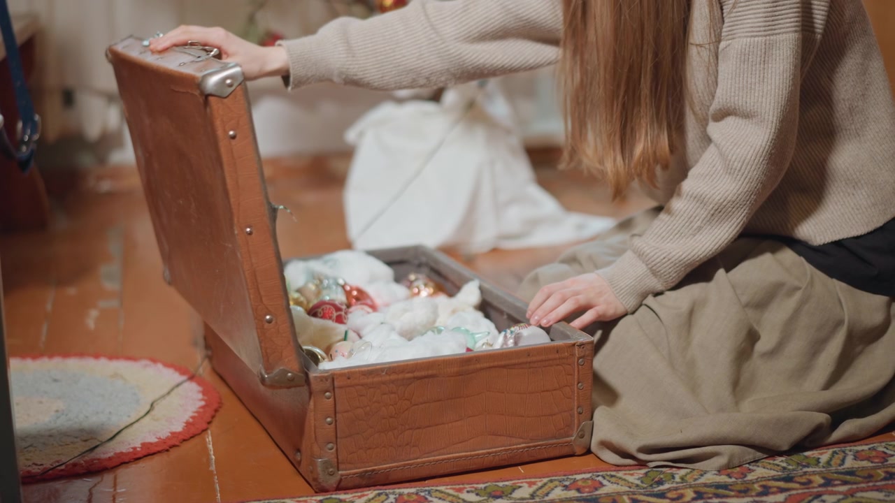 Nainen avaa vintage-arkun, joka on täynnä koristeita ja vanua. Kodikas olohuone, villapaita, kuviollinen matto, puulattia. Hellä arkistoijan hetki, kun hän paljastaa perintökoristeita.