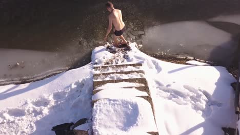 man standing on snow covered steps leading to frozen lake with small area of broken ice