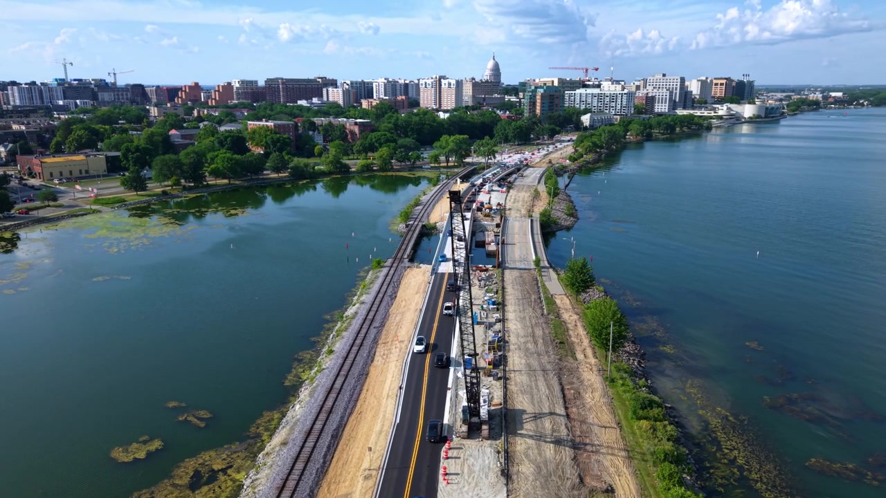 Luftfoto med drone, der flyver over en dæmningsvej og jernbane under opførelse ved en sø, med en kran, trafik og Madison, Wisconsin byens skyline i baggrunden på en solrig dag.