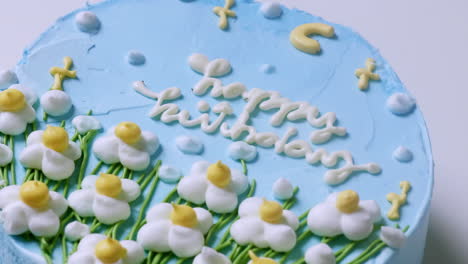 a close-up shot of a birthday cake with blue creamy frosting and decorated with white and yellow flowers, with green trimmings