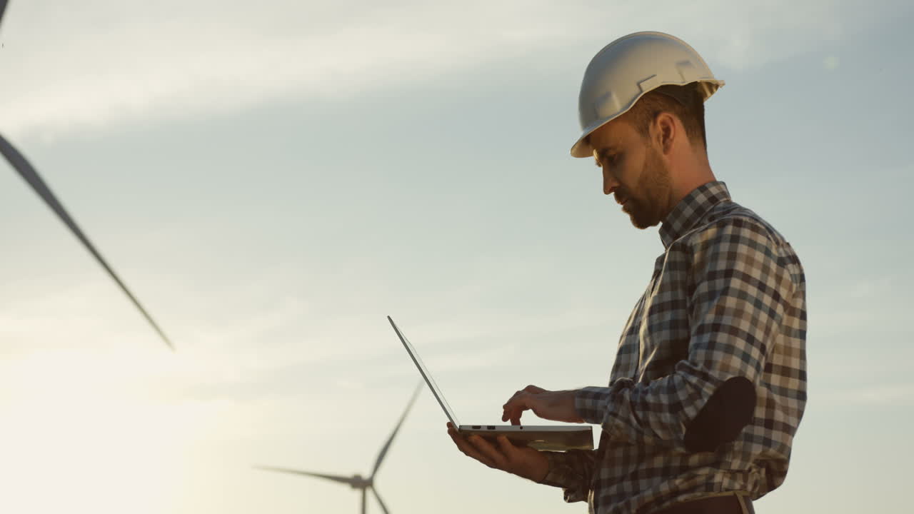 Side view of Caucasian man engineer wearing a helmet using laptop while checking operation of the windmill tourbines