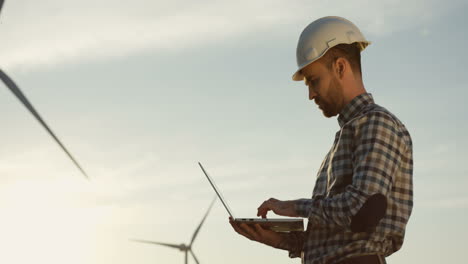 vista lateral del ingeniero caucásico que usa un casco usando una computadora portátil mientras verifica el funcionamiento de las turbinas del molino de viento