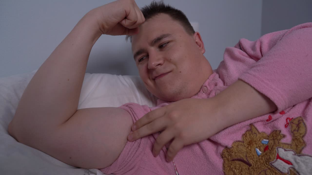 joven caucásico sobreponderado con cabello corto con pijama rosada divertida, acostado en la cama, muestra sus músculos en los bíceps, se besa allí y asume. concepto de humor de deportes estilo de vida no saludable.