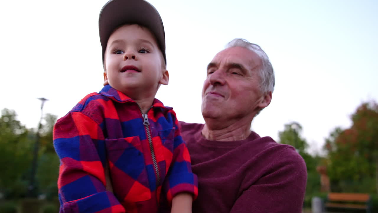 Grey-haired Caucasian man carrying a cute baby boy wearing checkered shirt and cap. Grandpa and grandson are walking outdoors. Close up.