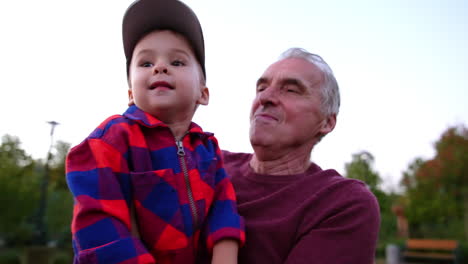 grey-haired caucasian man carrying a cute baby boy wearing checkered shirt and cap. grandpa and grandson are walking outdoors. close up.
