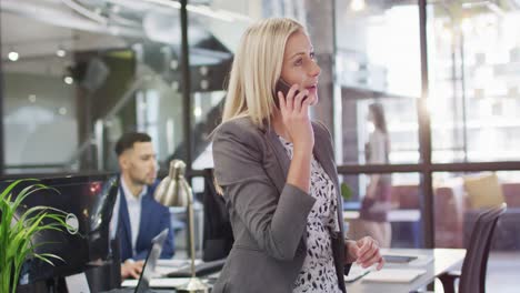 Empresaria-Caucásica-Hablando-Por-Teléfono-Inteligente-Con-Colegas-Que-Trabajan-En-Segundo-Plano.