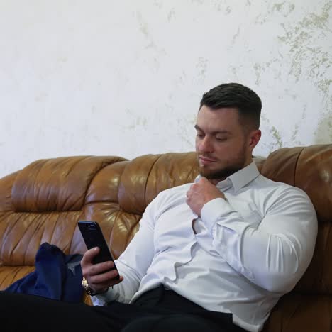 Handsome man on couch with a mobile phone. Young businessman sitting on a sofa in a hotel and unbuttoning his white shirt while viewing in the phone