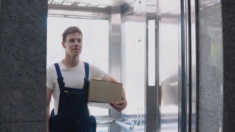 a delivery worker in a white t-shirt and blue jumpsuit comes out of the lift holding a box in his hand. delivery of goods to the office