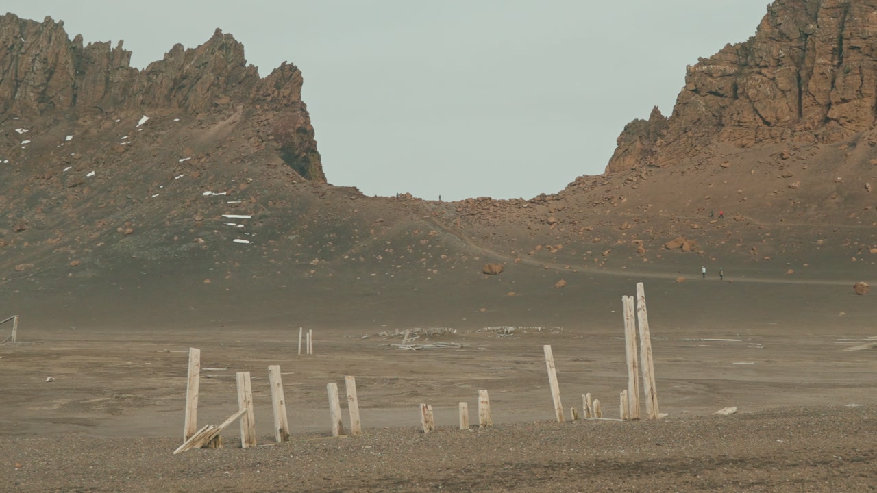 Weathered wooden posts, remnants of historic pier, stand across barren dried lake bed at Whalers Bay, Deception Island, framed by jagged volcanic peaks and tourists far in backdrop